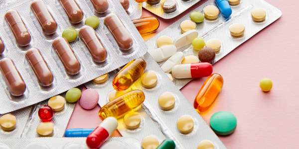 Mehrere Blisterpackungen mit Tabletten und Kapseln liegen auf einer rosa Fläche, dazwischen lose bunte Pillen. Darstellung kann auf die Kontrolle von Neben- und Wechselwirkungen verschiedener Medikamente hinweisen.